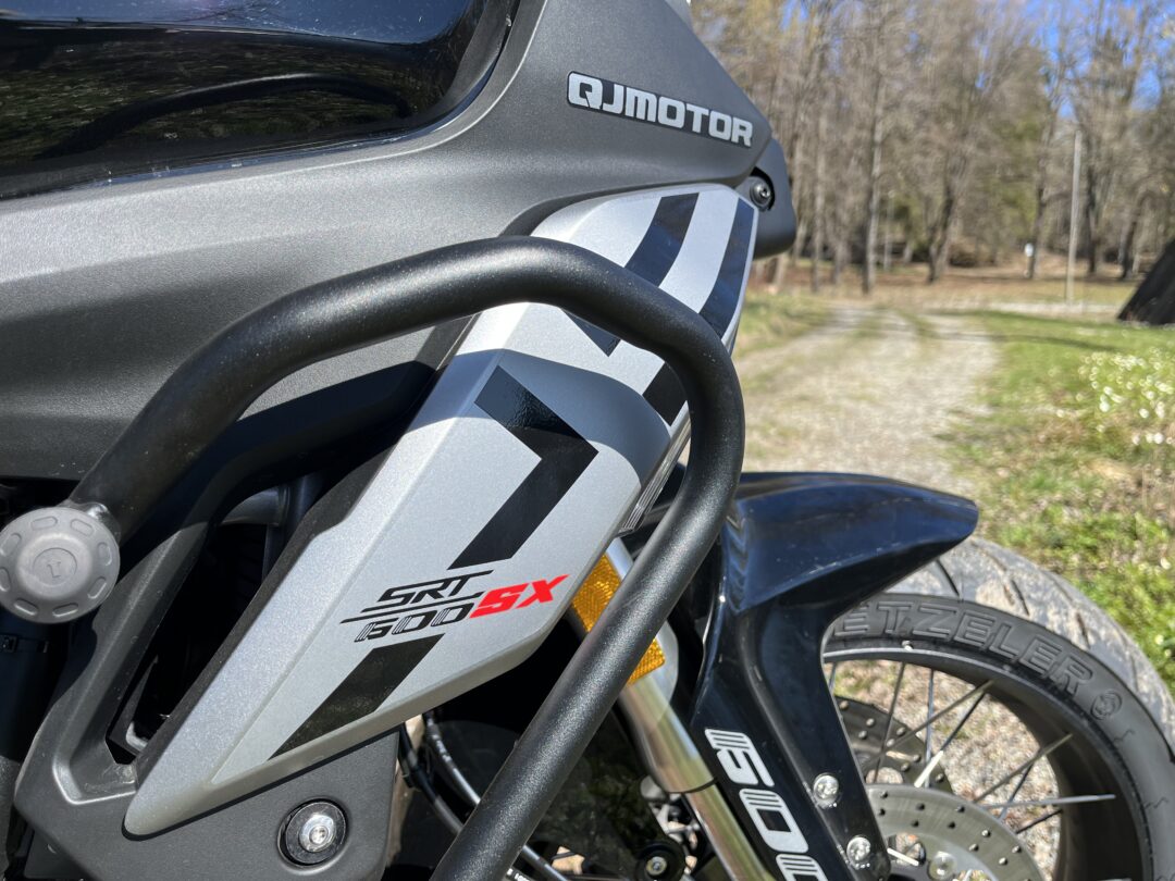 Close-up of a QJ Motor motorcycle front fairing with gray graphic, black crash bar, and partial front tire outdoors.