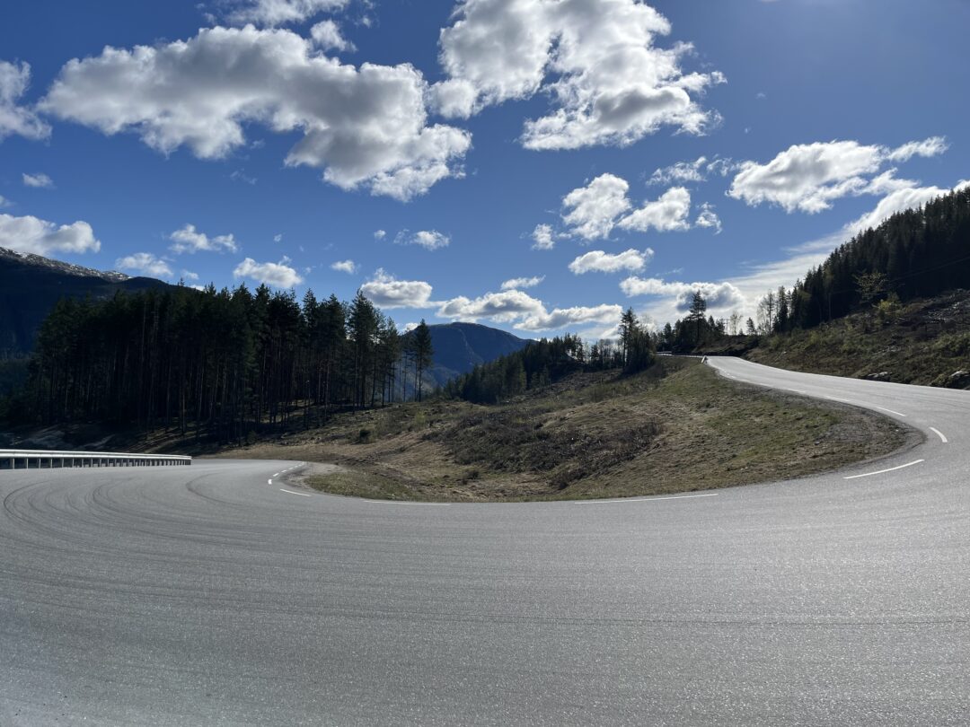 Winding mountain road curves through a forest under a bright blue sky with white clouds.