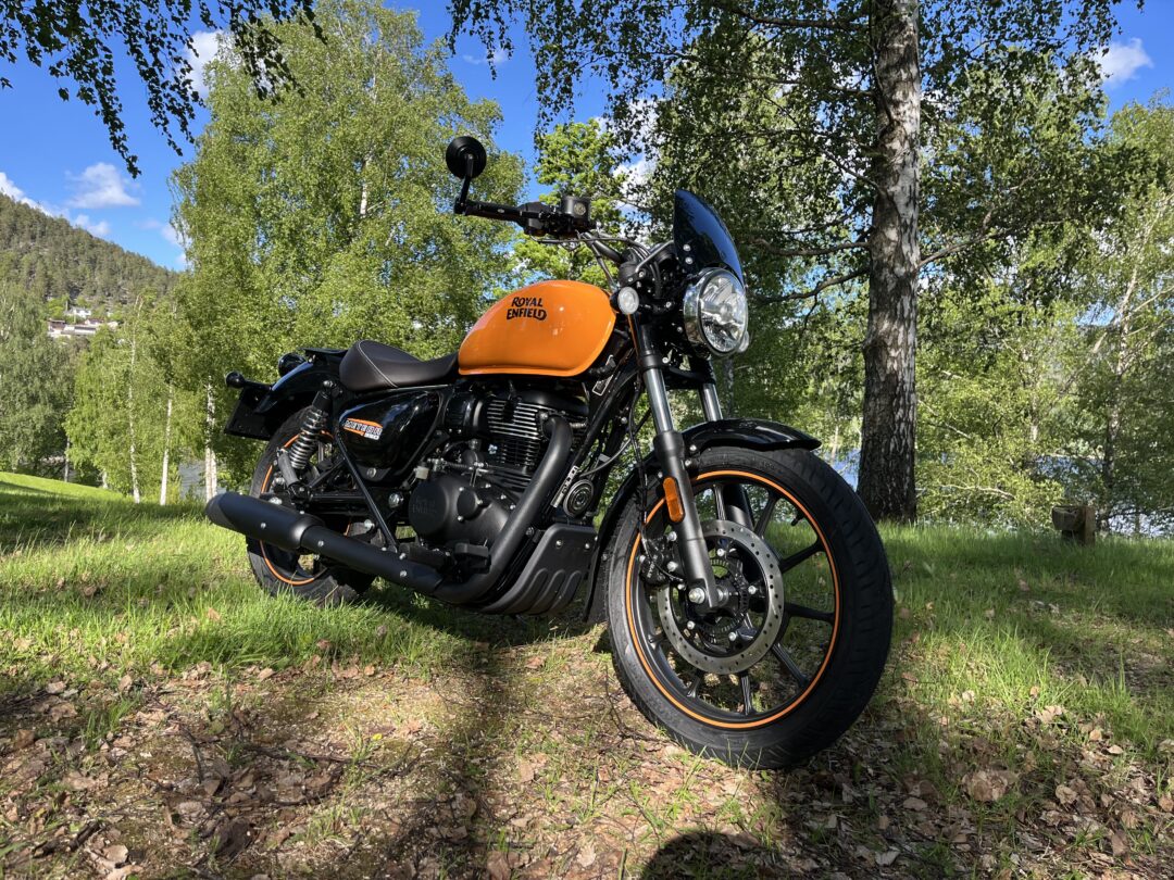 Orange Royal Enfield motorcycle parked on grass with trees and blue sky in a park setting.
