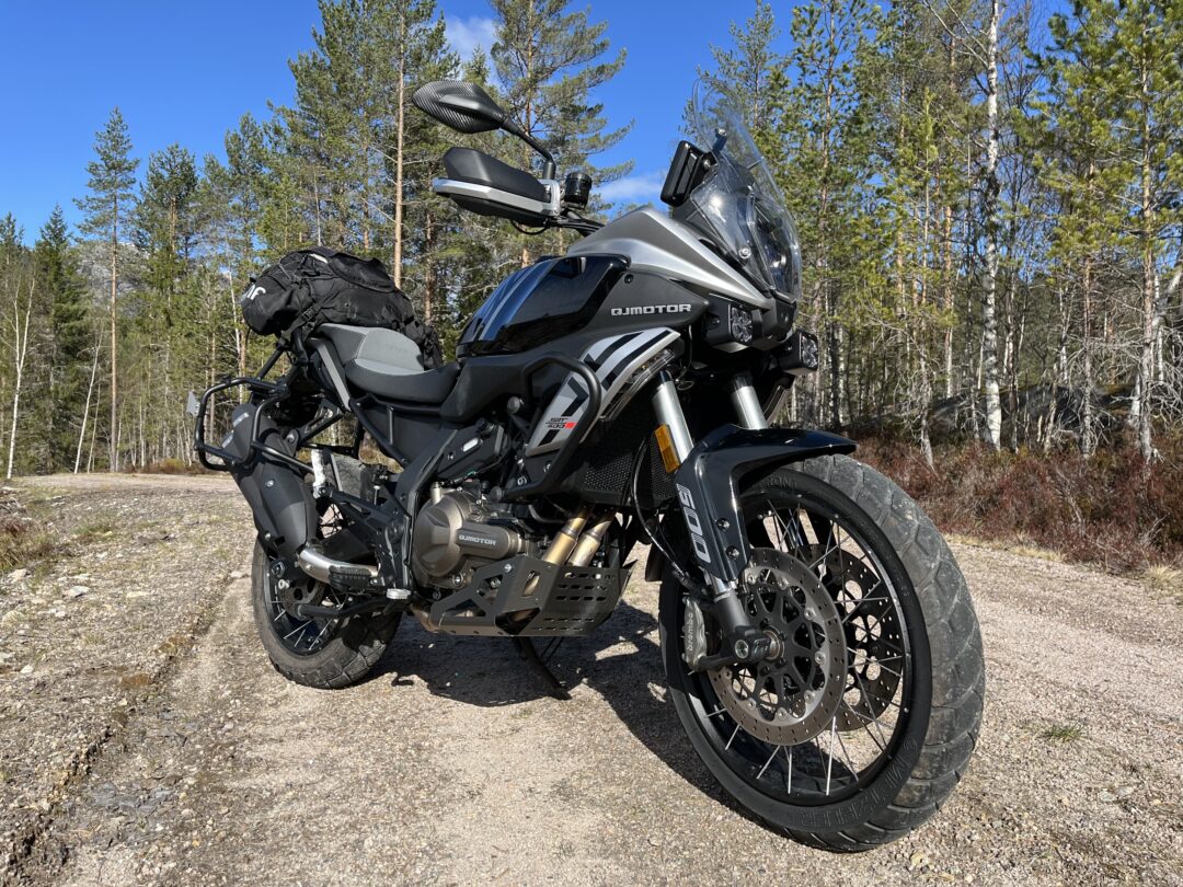 Adventure motorcycle parked on a gravel road with forest trees in the background, loaded with side panniers and a handlebar bag for a long ride.