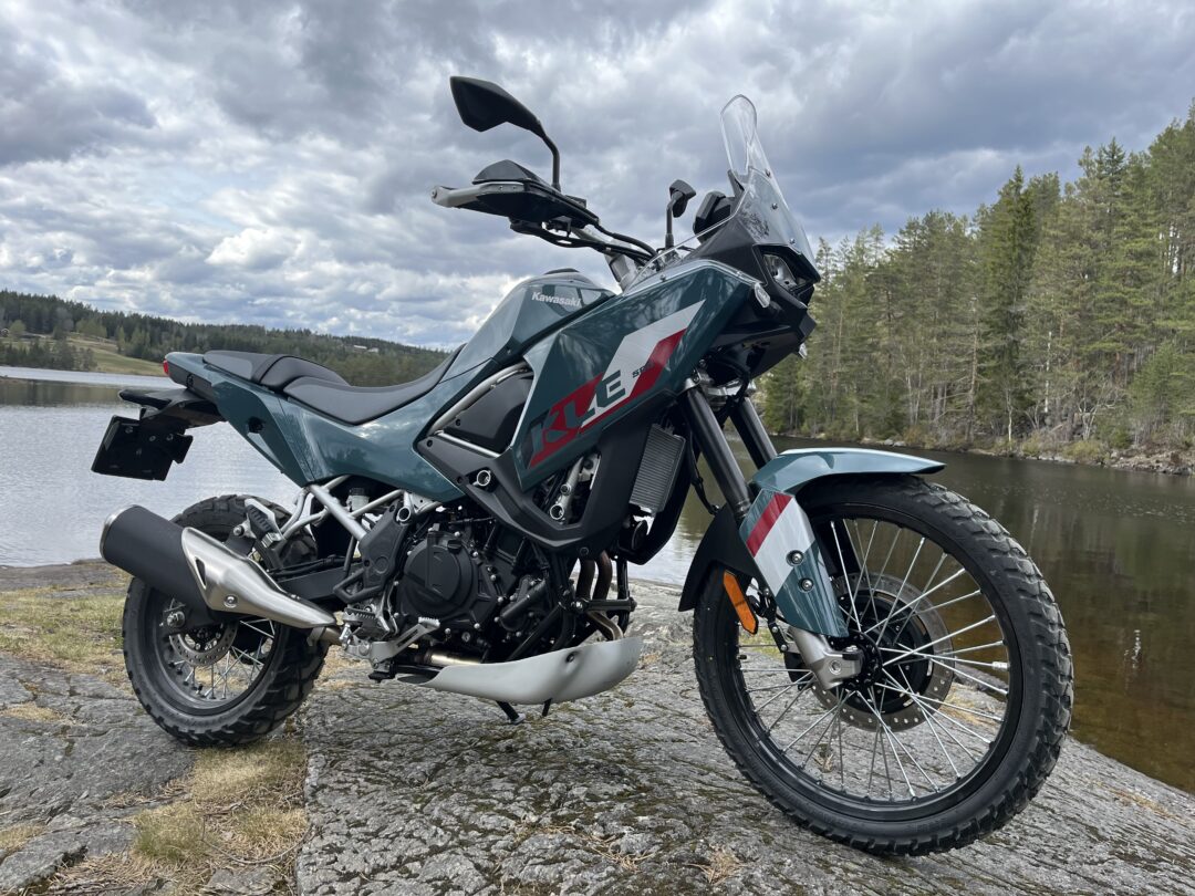 Kawasaki adventure motorcycle parked on a rocky lakeside shore with trees and cloudy sky in the background.