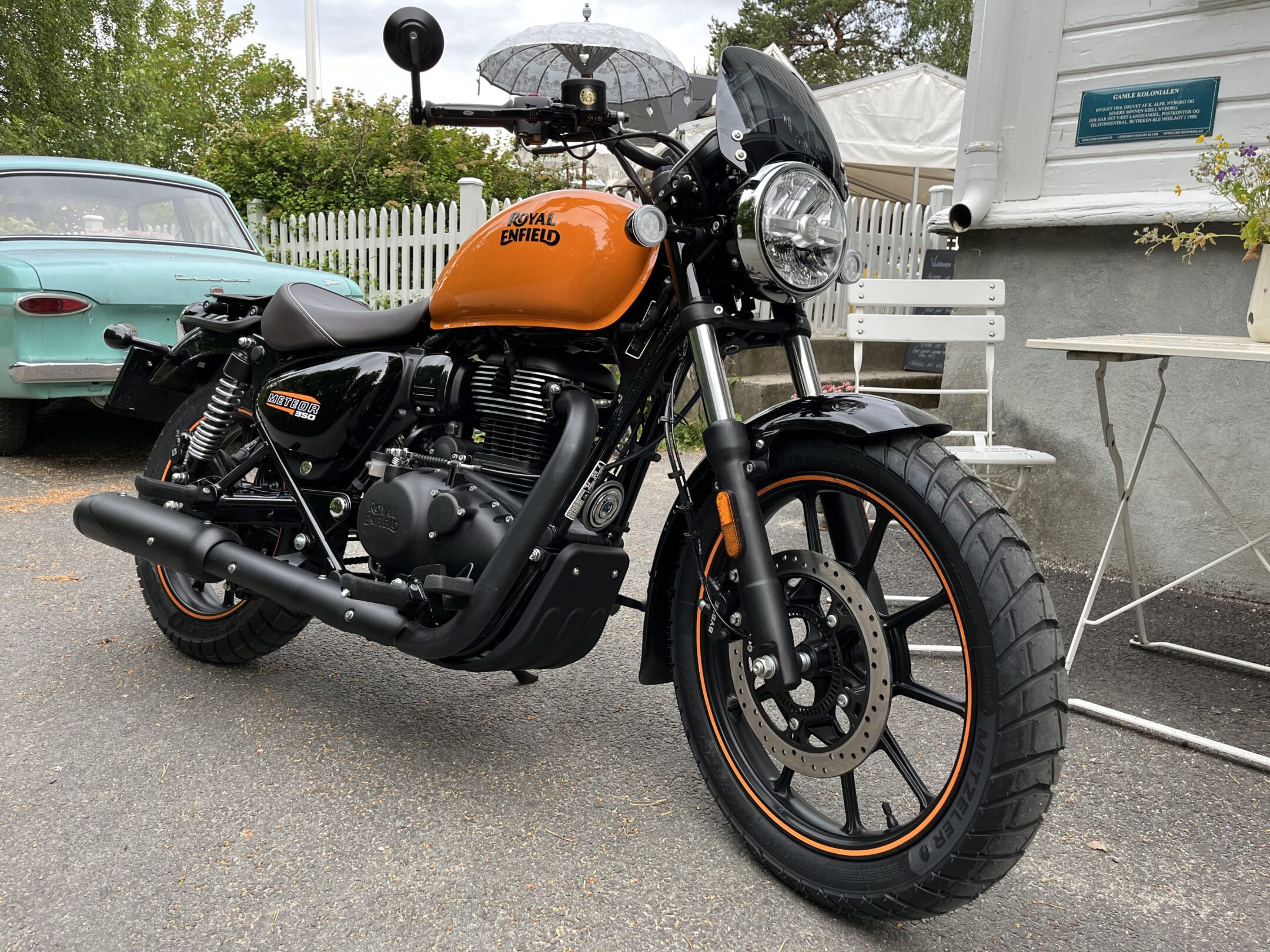Orange Royal Enfield motorcycle in foreground with a turquoise vintage car and white picket fence in the background, outdoor parking scene.