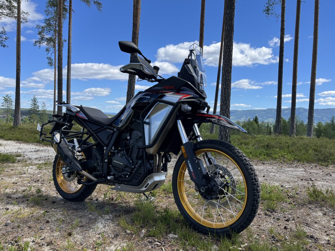 Adventure motorcycle with yellow rims parked on a dirt path among tall pine trees under a bright blue sky and distant hills in the background