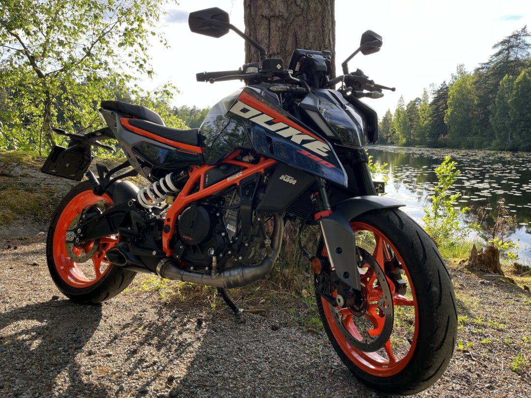 Sport motorcycle with bright orange rims parked beside a tree by a lake in a forested area, reflecting water in the background.