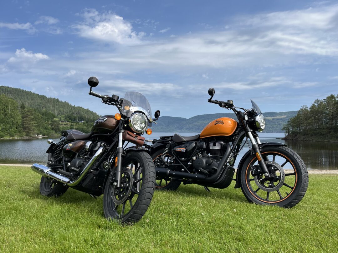 Two Royal Enfield motorcycles parked on a grassy lakeshore with hills and a blue sky behind.