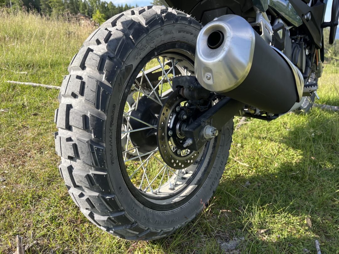 Close-up of a knobby off-road motorcycle rear wheel with disc brake and silver exhaust, on a grassy field in daylight.