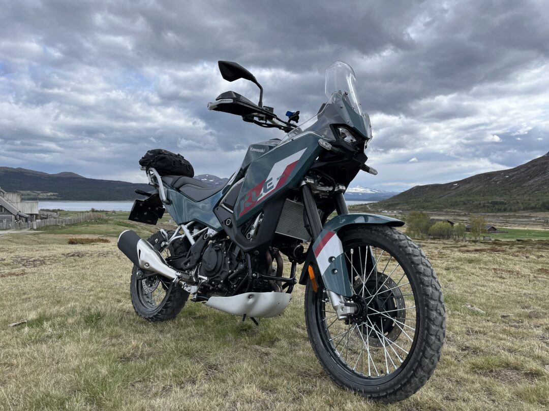 Blue Kawasaki adventure motorcycle with side luggage on grassy field, cloudy sky and distant hills in a rural landscape