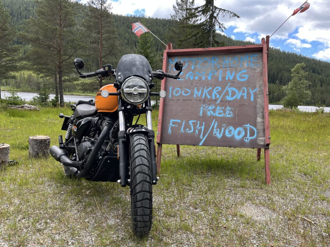 Motorcycle in foreground beside a wooden sign with blue lettering, a Norwegian flag atop, with trees and lake in the background.
