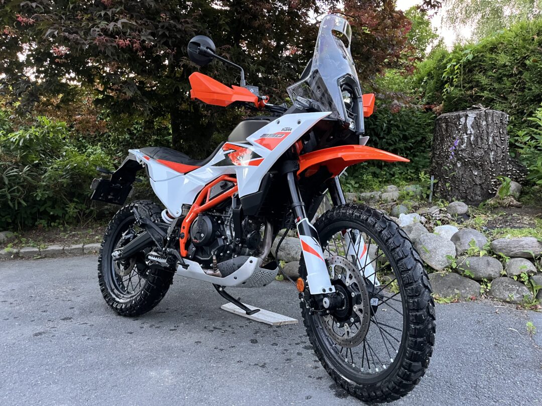 Orange and white KTM 390 Adventure motorcycle parked on a driveway with trees in the background, side view.