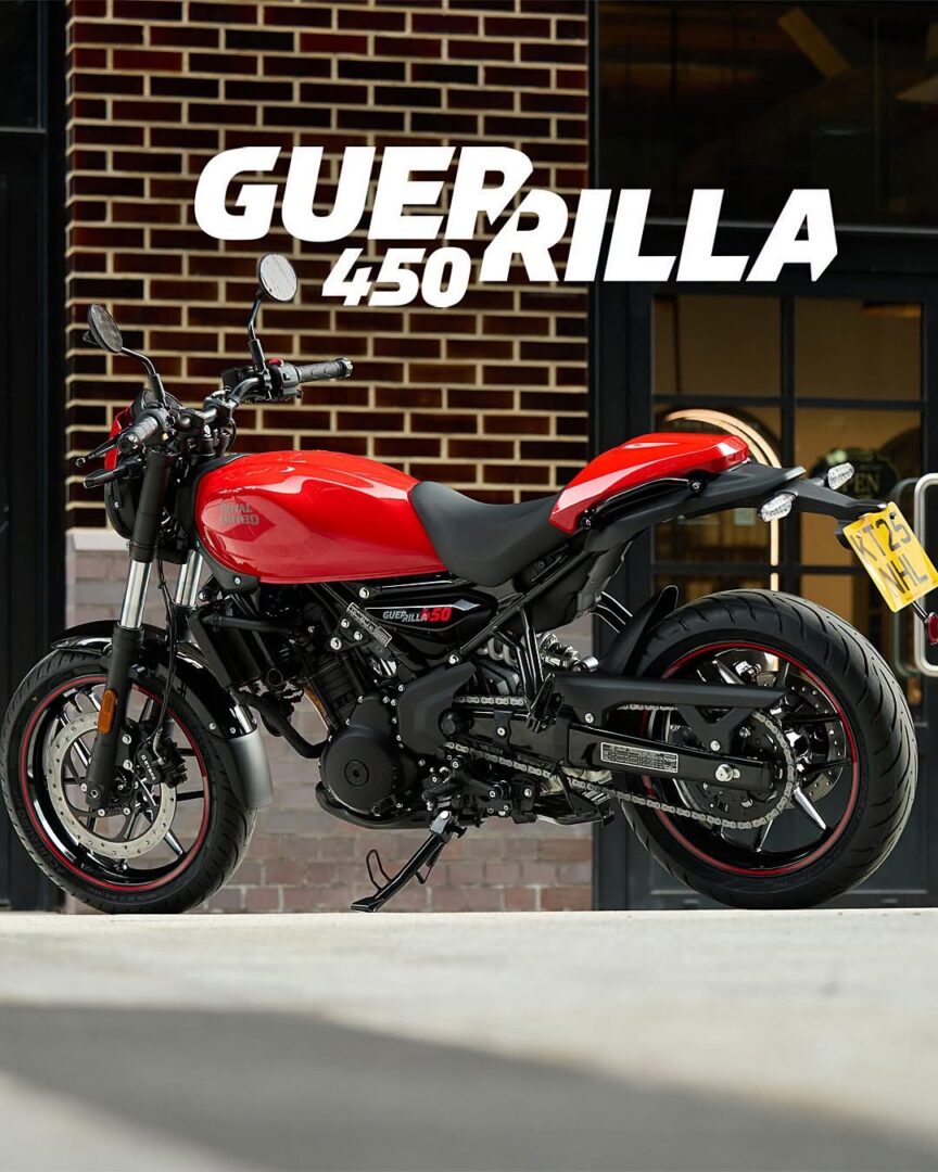 Red Guerilla 450 motorcycle parked outdoors beside a brick building, shown in a side profile with branding on the tank and prominent black frame.