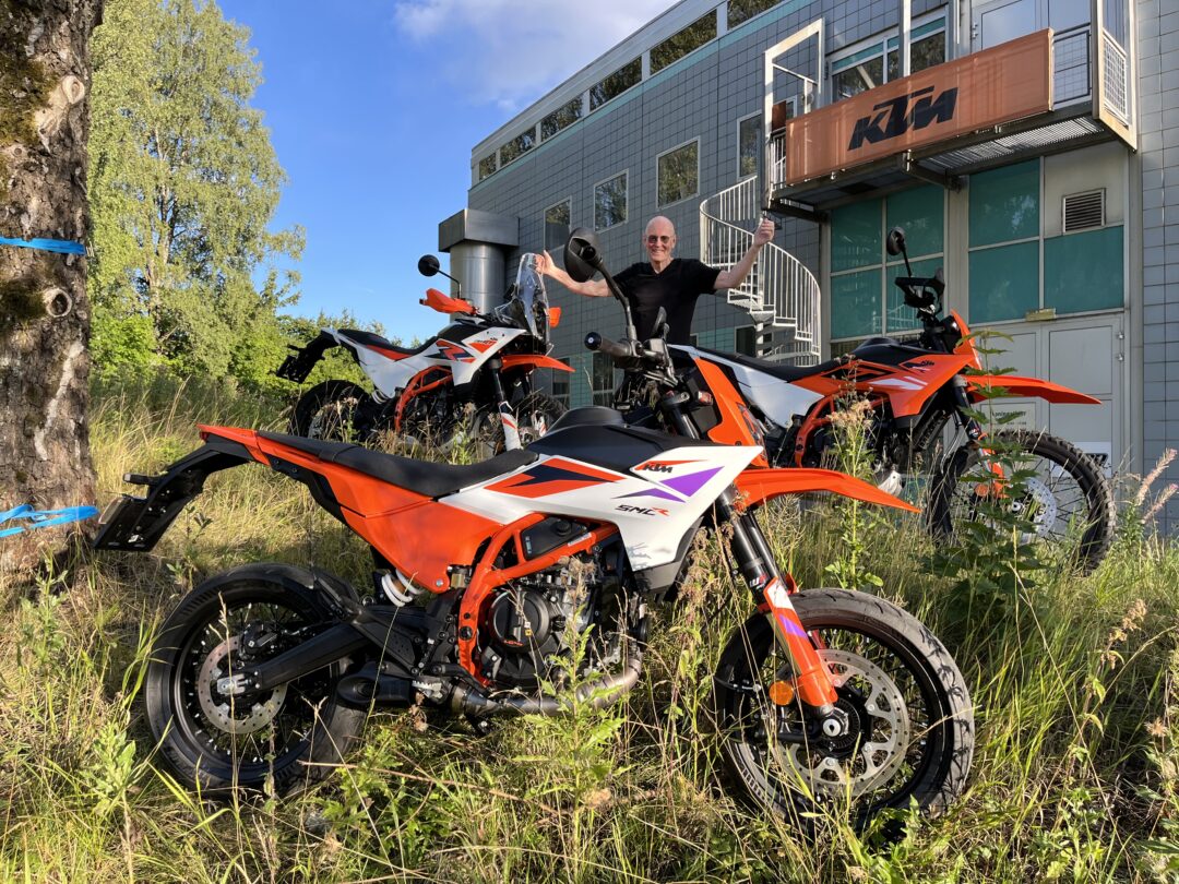 Man in a black shirt poses with orange KTM dirt bikes parked on grass outside a modern gray building with a KTM banner above.