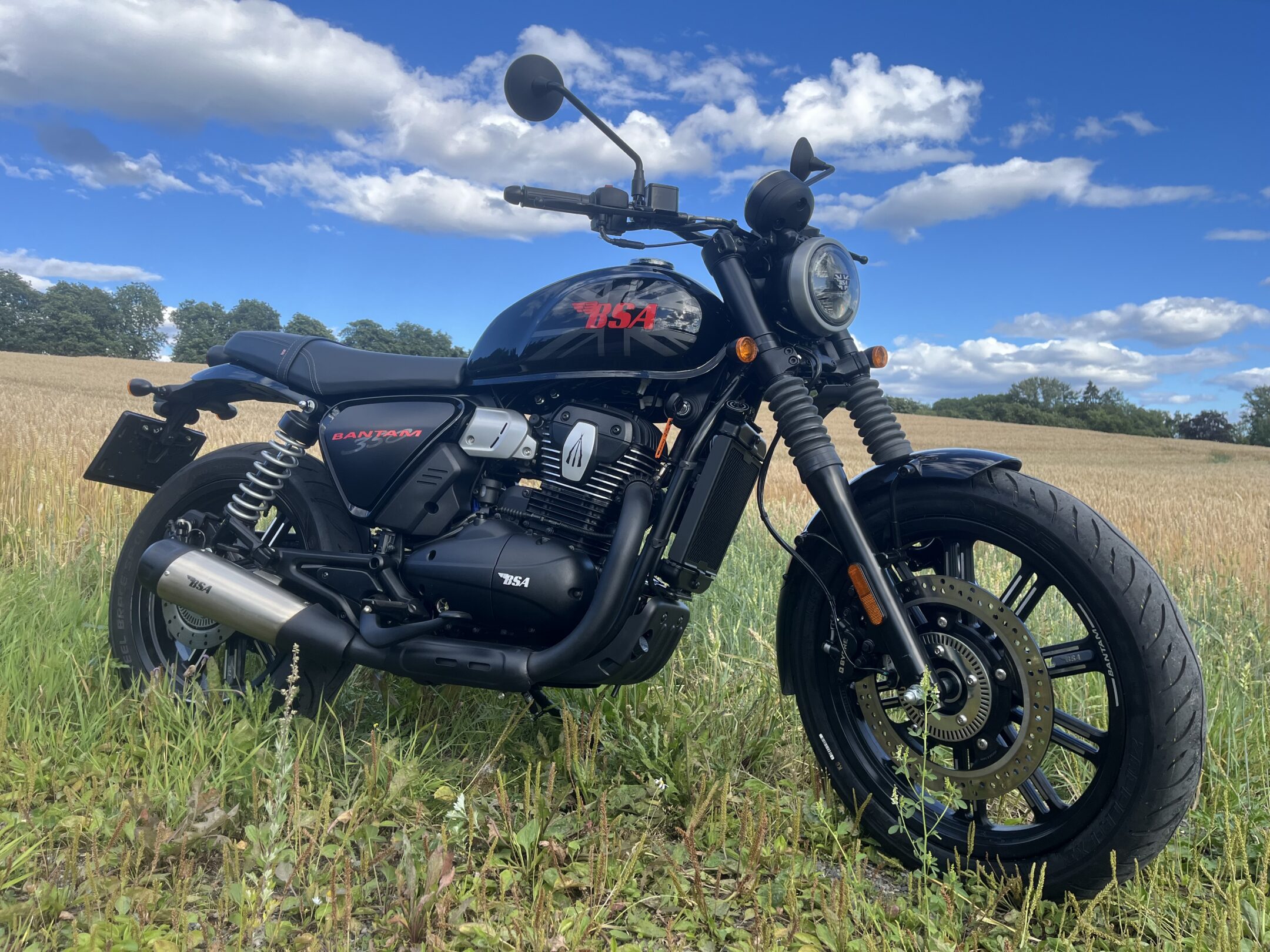 Black BSA Bantam motorcycle parked in a sunlit field with tall grass and a blue sky overhead.