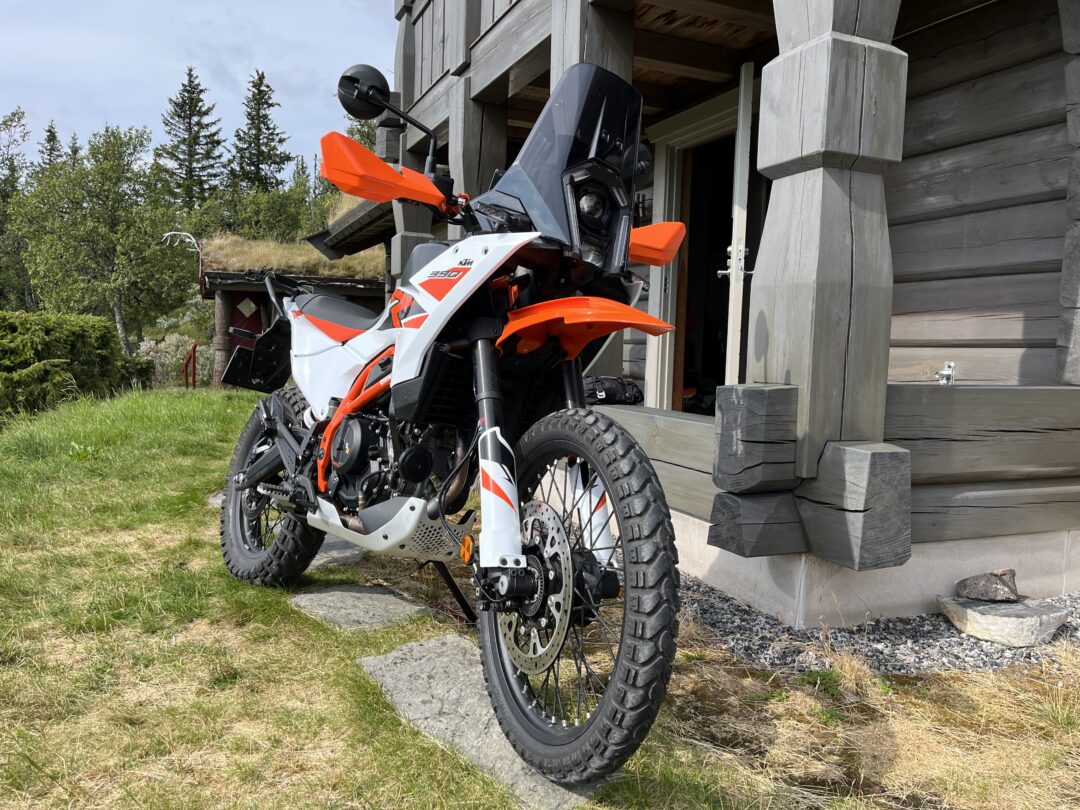 Orange and white KTM adventure motorcycle parked on a rocky driveway beside a wooden cabin, with a forested area in the background.