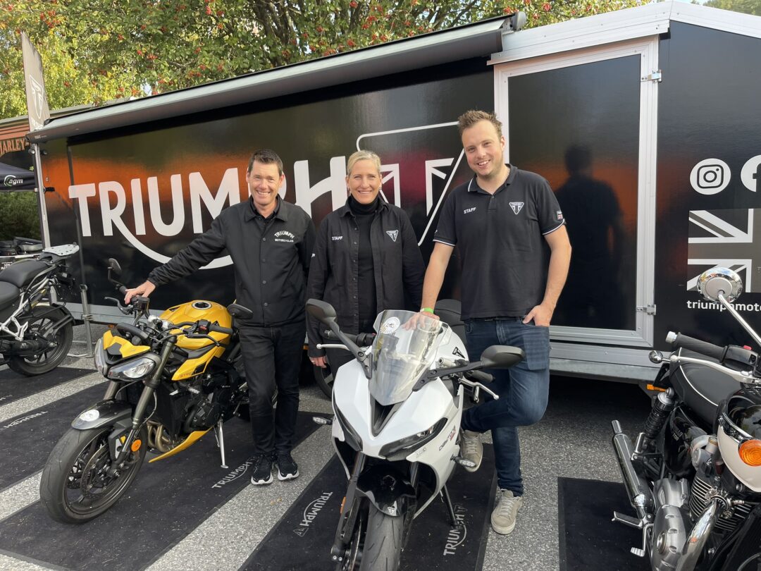 Three Triumph staff members pose with motorcycles in front of a black Triumph trailer at an outdoor event.